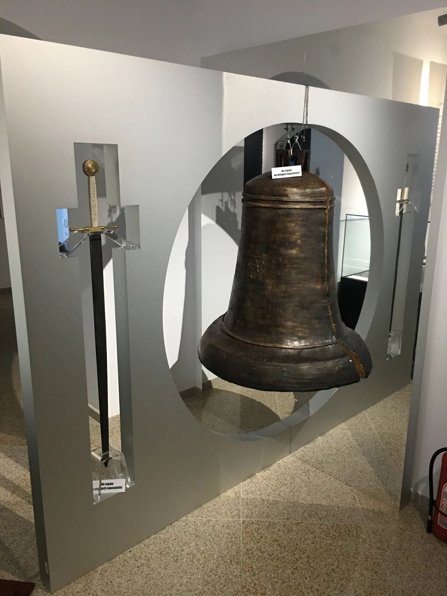 Panou expozițional cu un clopot și o sabie, în dreptul unei decupări ovale
