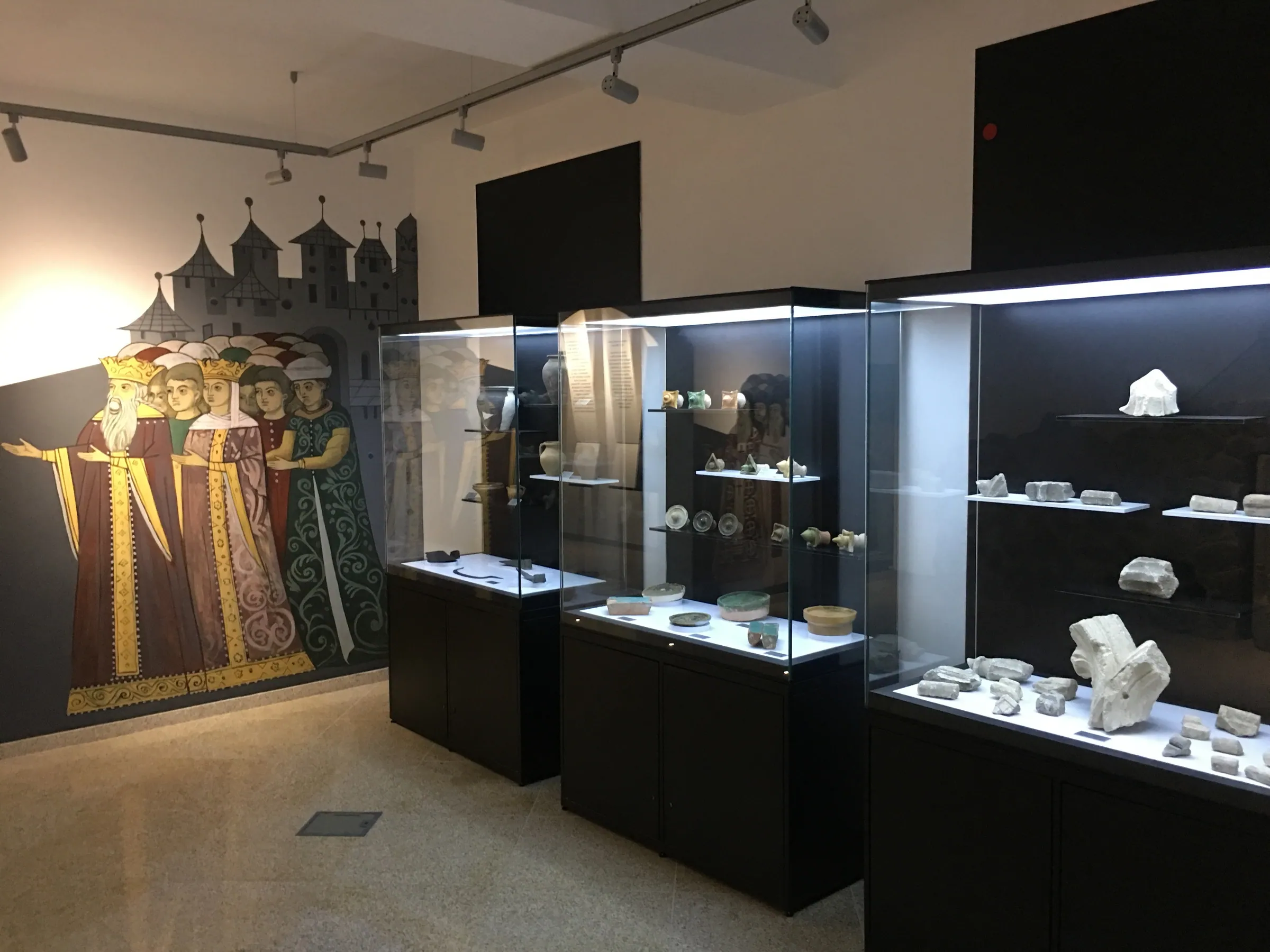 Vitrine cu fragmente de piatră și obiecte, lângă o pictură murală cu personaje medievale